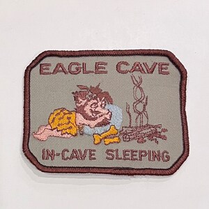 May include: Embroidered patch with the text "EAGLE CAVE" and "IN-CAVE SLEEPING". The patch features a cartoon caveman sleeping near a fire and a bone. The patch has a brown border and a gray background.
