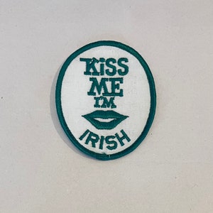 May include: A white round patch with green embroidered text that reads "Kiss Me I'm Irish" and a green embroidered lip design.