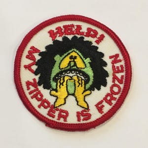 May include: A white embroidered patch with a red border. The patch features a green and black cartoon character with the text "My Zipper Is Frozen" around the character.