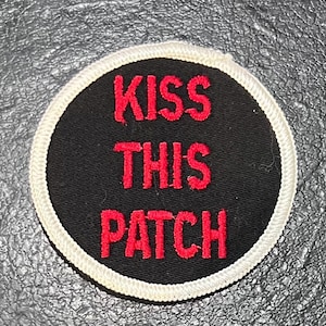May include: A black round patch with a white border. The text "KISS THIS PATCH" is embroidered in red.