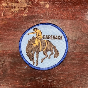 Peut inclure: Un écusson brodé bleu et blanc avec un cow-boy chevauchant un cheval brun. Le texte "BAREBACK" est brodé en brun sur l'écusson.
