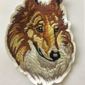 May include: Embroidered patch depicting a Collie dog's head. The patch features brown, gold, and grey thread, with a white border. The design is intricately stitched, capturing the dog's features. A decorative item for clothing or accessories.