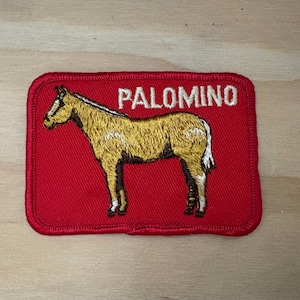 May include: A rectangular embroidered patch with a red background featuring a golden-brown horse standing in profile. The word PALOMINO is stitched in white capital letters above the horse. The patch has a thick red stitched border.