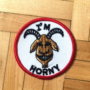 May include: A white round patch with a red border. The patch features a cartoon goat with horns and a toothy grin. The text "I'M HORNY" is embroidered in black around the goat.
