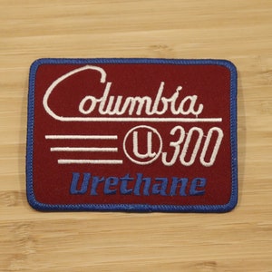 Pode incluir: Um remendo bordado vermelho com texto branco que diz "Columbia U 300 Urethane". O remendo tem uma borda azul.