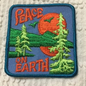 May include: A blue embroidered patch with a sunset scene and the text "Peace on Earth". The patch features two green trees, a red sun, and a flying bird.