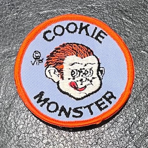 May include: A blue and orange embroidered patch with the text "COOKIE MONSTER" and a cartoon image of Cookie Monster with a red tongue sticking out.