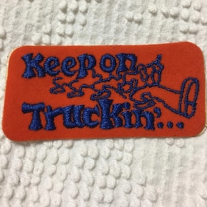 May include: An orange rectangular patch with the words "Keep on Truckin'" embroidered in blue. The patch features a stylized illustration of a truck. The background is a white, textured fabric.