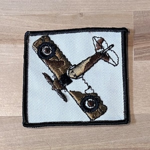 May include: A brown and white embroidered patch of a vintage biplane airplane with a black border.