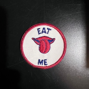 May include: A white circular patch with a red border. The patch has blue embroidery that reads "EAT ME" and a red embroidered tongue sticking out.
