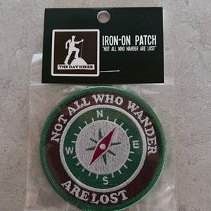 May include: Circular iron-on patch featuring a compass design and the words "NOT ALL WHO WANDER ARE LOST." The patch has a green and brown border, with a white compass rose and red accents. A black tag above reads "IRON-ON PATCH."