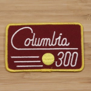 Op de afbeelding: Een geborduurde patch in bordeauxrood met een witte rand en een gele cirkel. De patch heeft de tekst "Columbia 300" in witte cursieve letters.