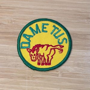 May include: A yellow and green circular patch with the word "Dame Tus" written in a green cursive font around the edge. A red cartoon donkey is in the center of the patch.