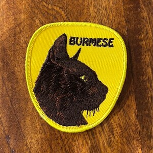 Op de afbeelding: Gele geborduurde patch met een zwart kattenhoofd en het woord "Burmese" in zwart.