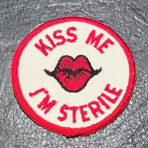 May include: A white round patch with a red border. The patch has red text that says "Kiss Me I'm Sterile" and a red lip with a black outline.