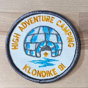 May include: A round, grey embroidered patch with the text "HIGH ADVENTURE CAMPING KLONDIKE 91" in gold lettering. The patch features a white and blue igloo with a star in the entrance.