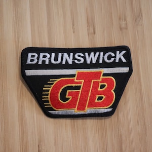 May include: Black and white embroidered patch with the word "BRUNSWICK" at the top. Below, the letters "GTB" are in red with a yellow outline. The patch has a black border and a silver stripe.
