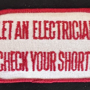 May include: Rectangular white patch with red embroidered text: "LET AN ELECTRICIAN CHECK YOUR SHORTS." The patch has a red border and is set against a black background. A humorous item.