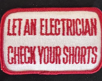 May include: Rectangular white patch with red embroidered text: "LET AN ELECTRICIAN CHECK YOUR SHORTS." The patch has a red border and is set against a black background. A humorous item.