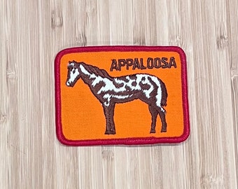 May include: An orange embroidered patch with a red border featuring a black and white Appaloosa horse. The text "APPALOOSA" is embroidered in brown above the horse.