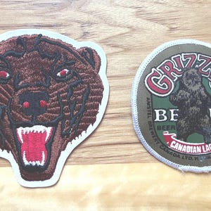 Op de afbeelding: Twee geborduurde patches met berenontwerpen. Eén patch is een bruine berenkop met een open mond en rode accenten. De andere is een ronde patch met een berensilhouet en de woorden "GRIZZLY BEER CANADIAN LAGER".