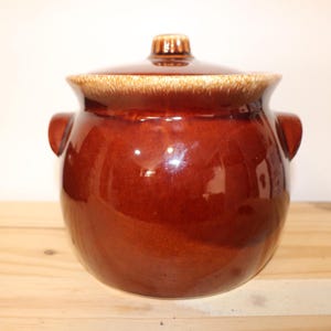 May include: A brown ceramic crock pot with a lid. The crock pot has two handles on the sides and a glazed finish.