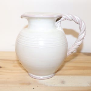 May include: White ceramic jug with a pink rope handle. The jug has a rounded body and a slightly flared lip.