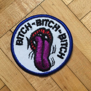 May include: Embroidered patch with a white background, a blue border, and the word "BITCH" repeated around the edge. The patch features a purple tongue with red and black details.