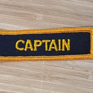 Peut inclure: Écusson rectangulaire bleu marine avec le mot "CAPTAIN" brodé en fil d'or. Le patch a une bordure dorée, probablement pour un uniforme ou un vêtement.