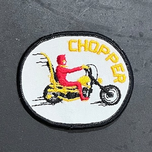May include: A white embroidered patch with a black outline featuring a red and yellow cartoon figure riding a yellow motorcycle with the word "CHOPPER" in yellow lettering.