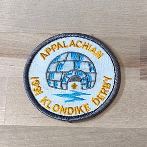 May include: A grey and white embroidered patch with the text "1991 Appalachian Klondike Derby" in gold lettering. The patch features a white igloo with blue accents and a gold star.