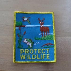 Op de afbeelding: Geborduurde patch met een blauwe achtergrond en gele rand. De patch toont een hert, een vis, een eend en de gele woorden "PROTECT WILDLIFE". De patch is rechthoekig.