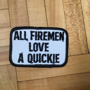 May include: White patch with black embroidered text that reads "ALL FIREMEN LOVE A QUICKIE".