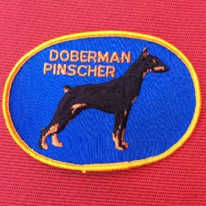 May include: A blue and yellow embroidered patch featuring a Doberman Pinscher dog silhouette and the text "Doberman Pinscher".