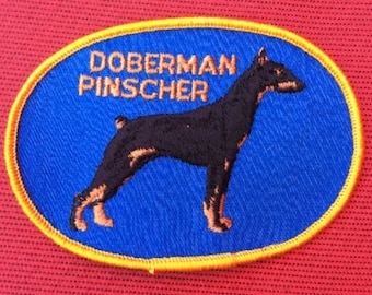 May include: A blue and yellow embroidered patch with a Doberman Pinscher dog silhouette and the text "Doberman Pinscher".