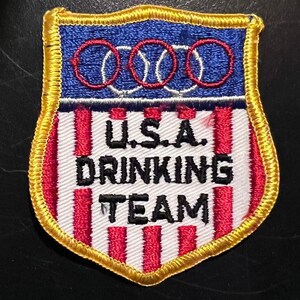 May include: A red, white, and blue embroidered patch with a gold border. The patch features three red circles with white baseballs inside, and the text "U.S.A. DRINKING TEAM".