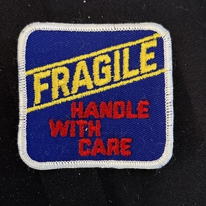 May include: A blue square patch with white stitching around the edges. The patch has yellow text that reads "FRAGILE" with a diagonal yellow line through it. Below the text is red text that reads "HANDLE WITH CARE".