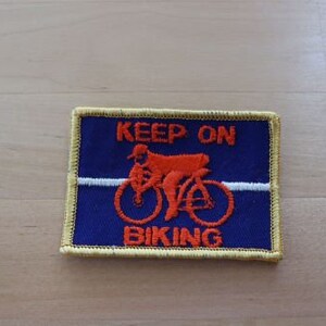 May include: A blue embroidered patch with yellow stitching. The patch features a red bicycle with a person riding it. The text "KEEP ON BIKING" is embroidered in white.