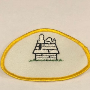 May include: A white embroidered patch with a yellow border featuring a cartoon dog on top of a doghouse. The dog is lying down with its head resting on its paws.