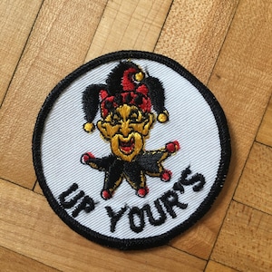 May include: A white round patch with a black border. The patch features a cartoon jester with a red and black hat, a yellow face, and a black and red costume. The text "UP YOURS" is written in black below the jester.