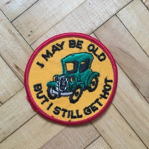 May include: A round, yellow embroidered patch with a red border. The patch features a green vintage car with the text "I may be old but I still get hot".