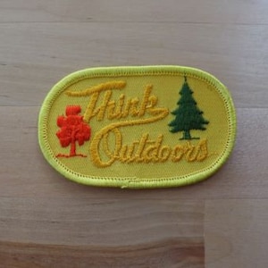 May include: Yellow oval-shaped patch with the text "Think Outdoors" in yellow embroidery. The patch features two embroidered trees, one green and one orange.