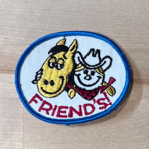 May include: A white oval embroidered patch with a blue border. The patch features a cartoon horse and cowboy with the text "FRIEND'S" in red.