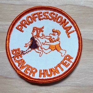 May include: A white round patch with an orange border. The patch has an embroidered image of two cartoon figures wrestling. The text "PROFESSIONAL BEAVER HUNTER" is embroidered around the edge of the patch.