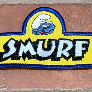 May include: Embroidered patch featuring the word "SMURF" in white, outlined in black, on a yellow background. A blue border surrounds the patch. Above the word is a blue Smurf character on a white background.
