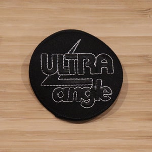 Può includere: Un cerotto circolare nero con cuciture bianche. Il cerotto presenta il testo "ULTRA angle" in un font stilizzato.