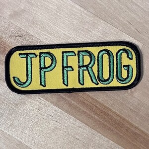 May include: A yellow and black rectangular patch with the text "JP FROG" in green embroidered letters.