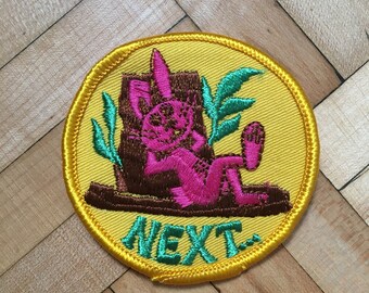 May include: Embroidered patch with a yellow background and a gold border. The patch features a pink rabbit lounging on a brown log with green leaves. The word "NEXT" is embroidered in green.