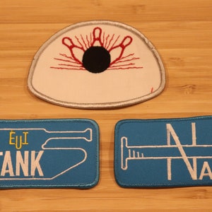 May include: Embroidered patches featuring a bowling pin design, a tank, and a nail. The bowling patch is white with red and black details. The tank and nail patches are blue with white text and designs. These patches are ideal for jackets, bags, or other fabric items.