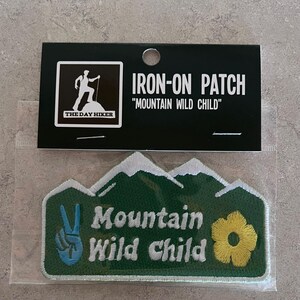 May include: An iron-on patch with the text "Mountain Wild Child" and a peace sign. The patch is green with white and yellow accents. The packaging includes the text "The Day Hiker".